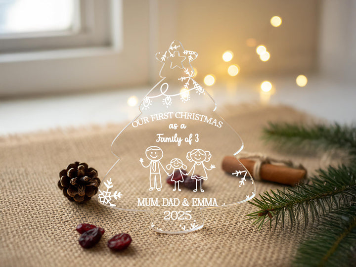 A clear glass ornament with a Christmas tree design, featuring a family of three and the text &quot;Our first Christmas as a Family of 3&quot; along with the names &quot;Mum, Dad & Emma&quot; and the year &quot;2025&quot;. The ornament is placed on a beige surface, surrounded by pine branches and pine cones, with Christmas lights visible in the background.