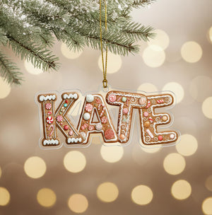 A clear ornament shaped like the word "KATE" made of gingerbread cookies, with various decorations and toppings, hanging from a branch of a pine tree.