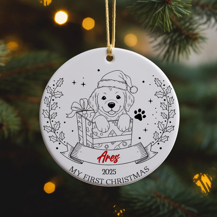 A white ornament with a black and white illustration of a dog wearing a Santa hat, sitting in a gift box, and surrounded by holly leaves and a ribbon.