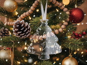 A close-up view of a Christmas tree adorned with various ornaments, including a clear ornament with the names "Isabella & David" and the year "2025" written on it.