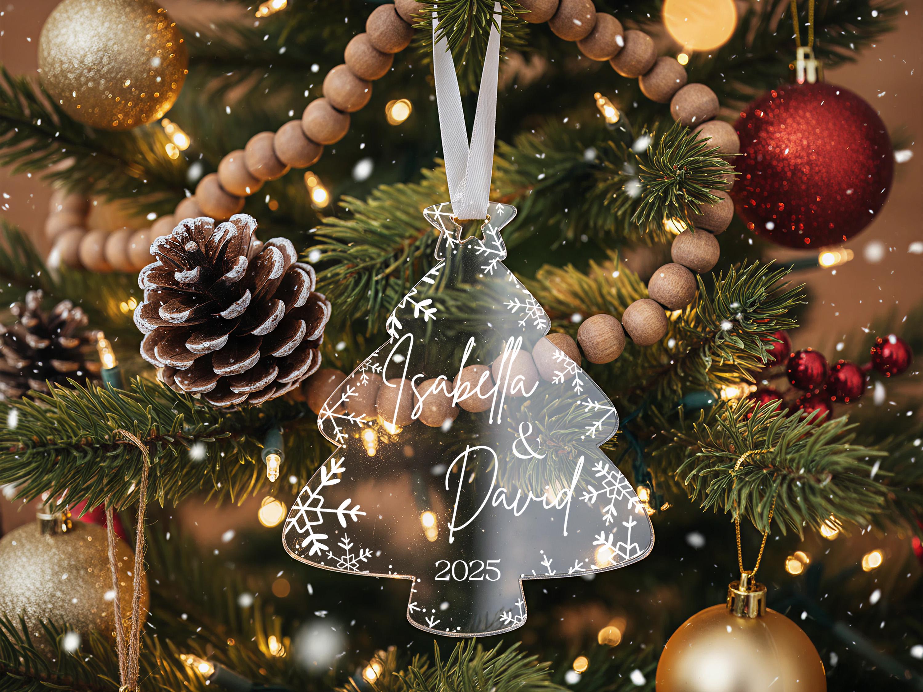 A close-up view of a Christmas tree adorned with various ornaments, including a clear ornament with the names "Isabella & David" and the year "2025" written on it.