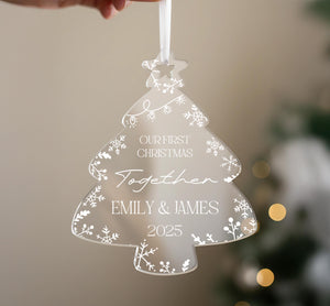 A hand holding a transparent ornament shaped like a Christmas tree with the text "Our first Christmas together" and "Together Emily & James" written on it.