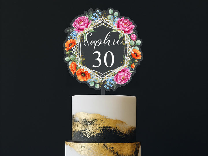 A cake topper with a floral design and the name &quot;Sophie&quot; written on it, placed on top of a two-tiered cake.