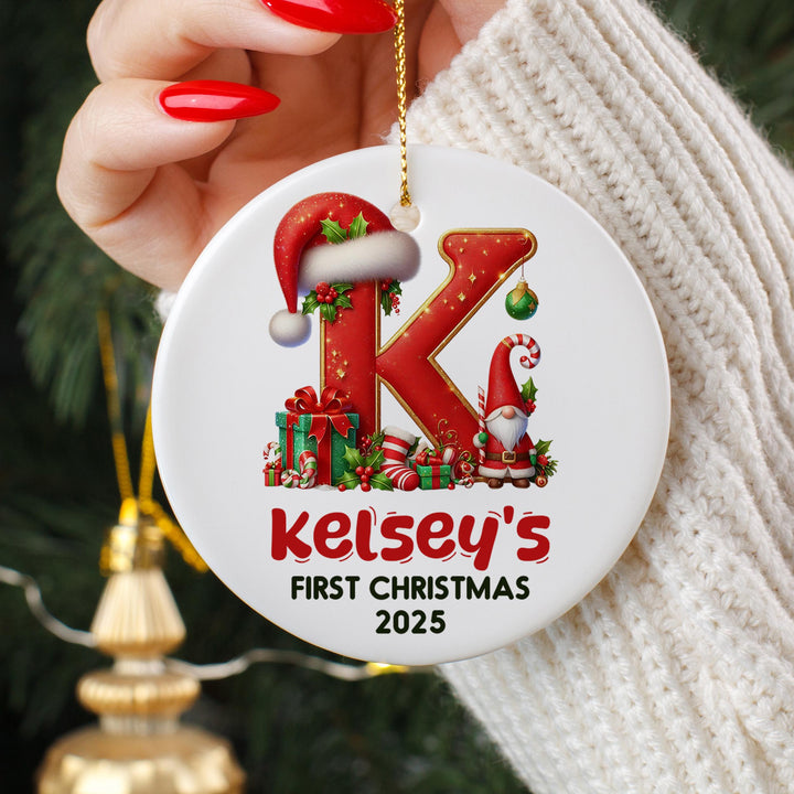 A person&#39;s hand holding a white ornament with a red letter &quot;K&quot; and various Christmas-themed decorations, including a Santa hat, presents, and a gnome.