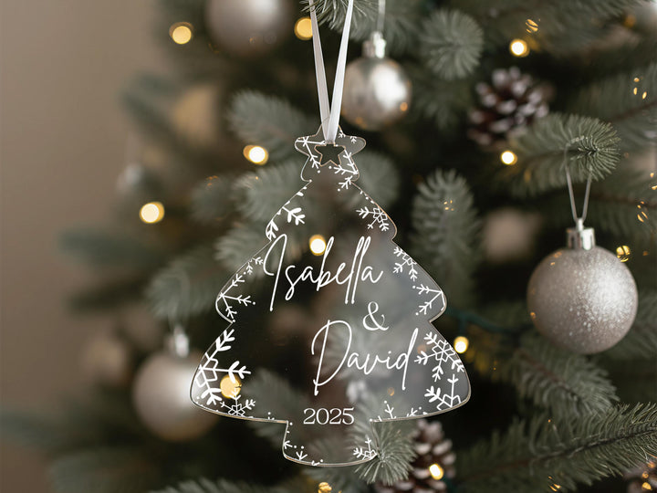 A clear ornament shaped like a Christmas tree with the names &quot;Isabella & David&quot; and the year &quot;2025&quot; written on it, hanging from a tree with other ornaments and lights visible in the background.