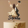 Personalised graduation cake topper featuring a female graduate in cap and gown, raising her arms in celebration, standing on a two-tier cake with gold floral decoration. Custom name and “Class of 2025” on gown.