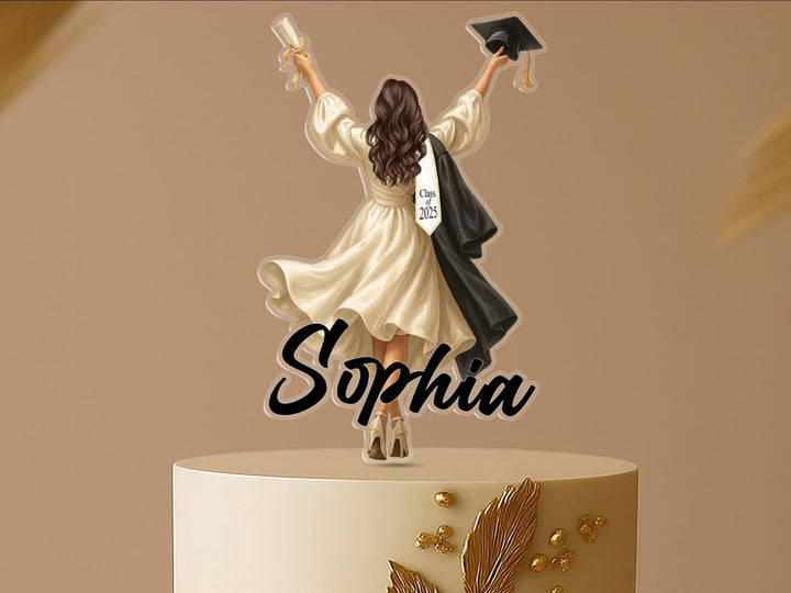Personalised graduation cake topper featuring a female graduate in cap and gown, raising her arms in celebration, standing on a two-tier cake with gold floral decoration. Custom name and “Class of 2025” on gown.
