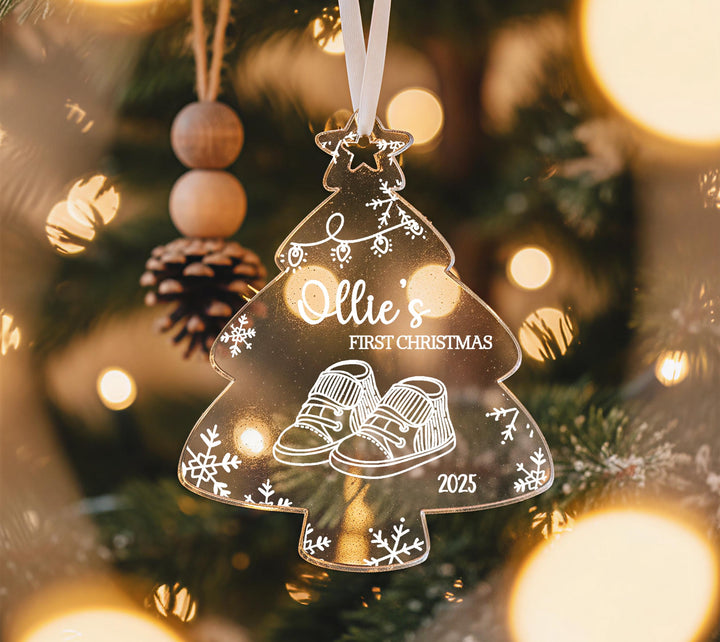 A clear ornament shaped like a Christmas tree with a snowflake design, hanging on a tree with lights visible in the background.