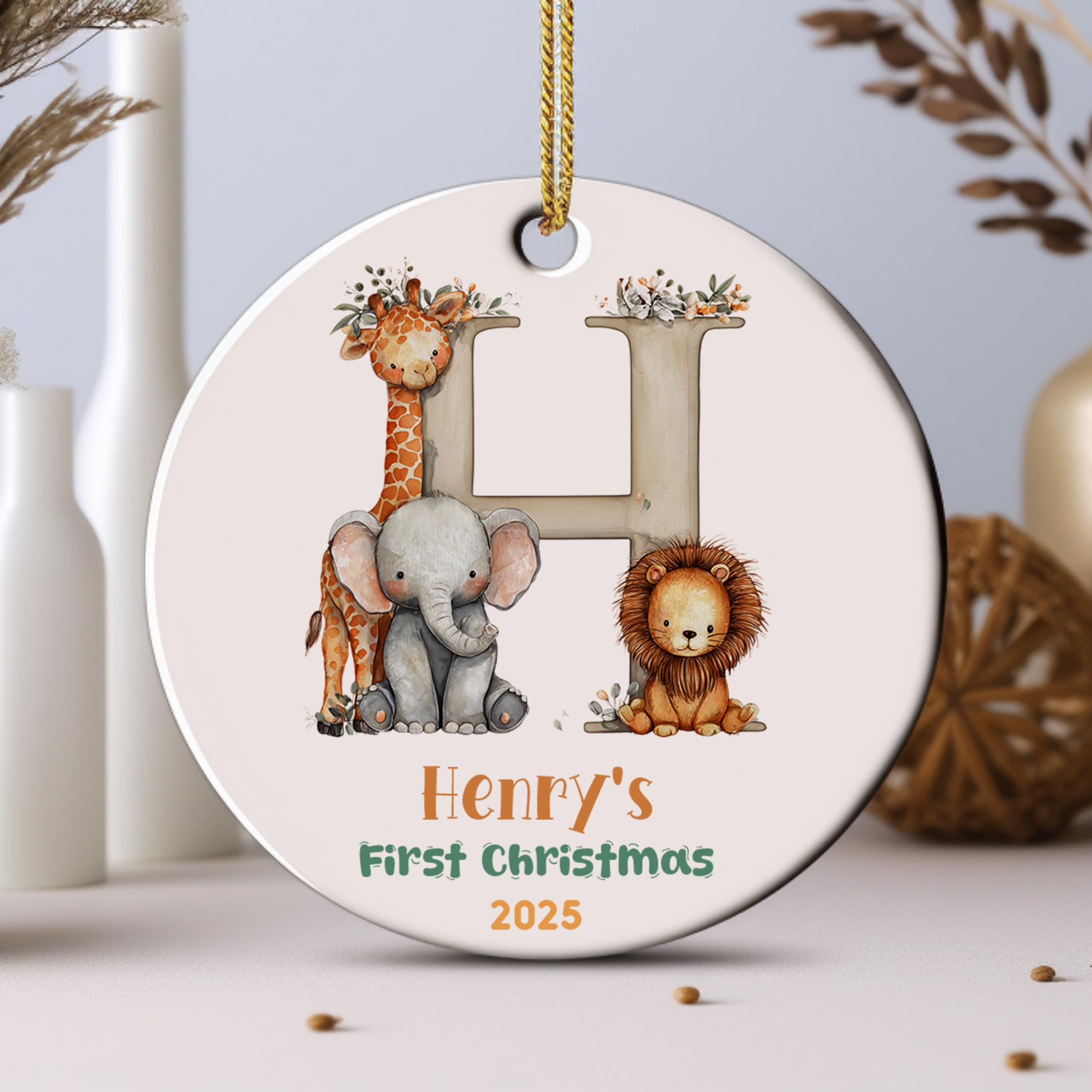 A decorative ornament featuring a large letter "H" with illustrations of a giraffe, elephant, and lion, along with the text "Henry's First Christmas 2025".