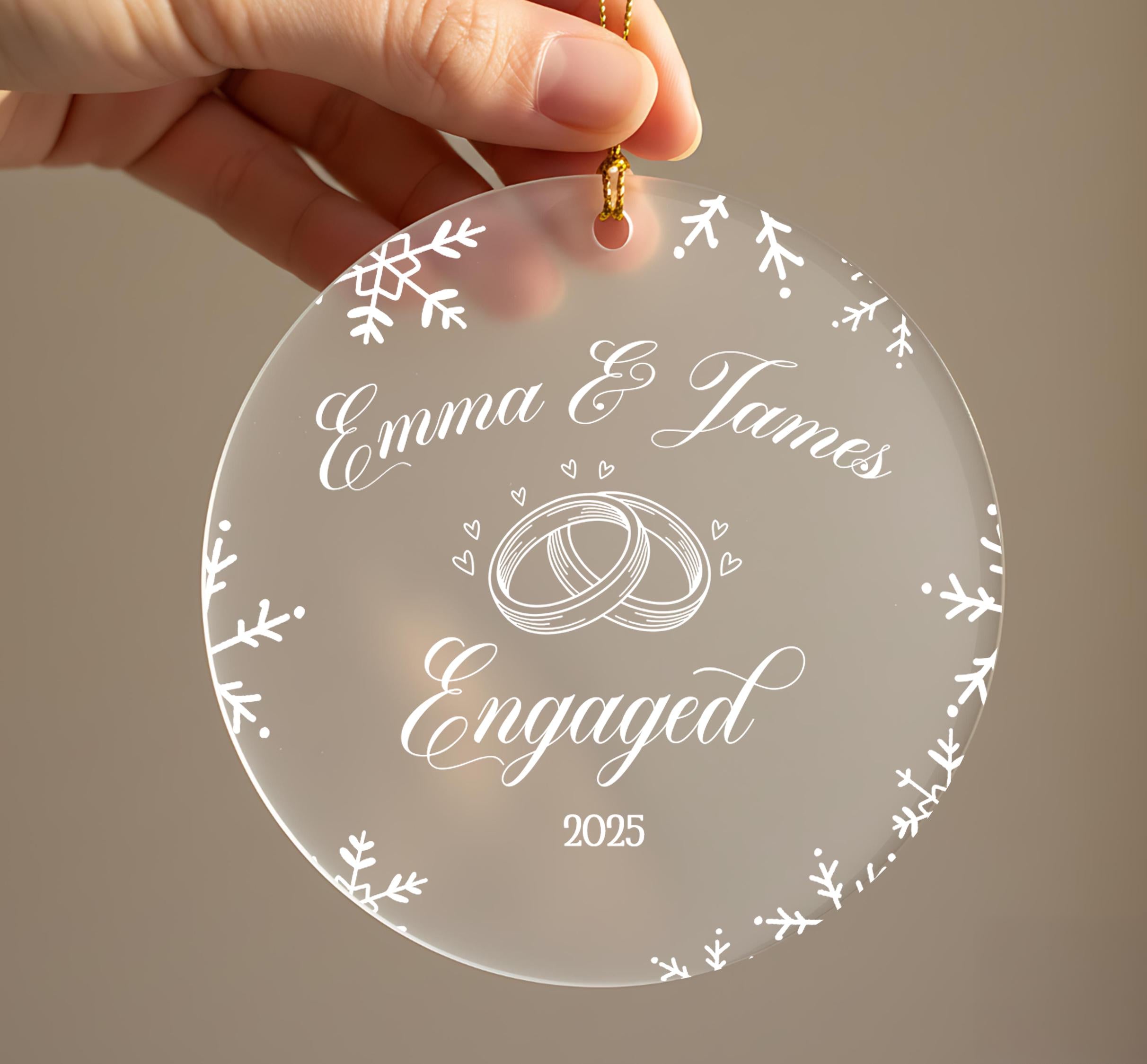 A hand holding a clear ornament with the text "Emma & James Engaged" and a graphic of a ring, surrounded by snowflakes.