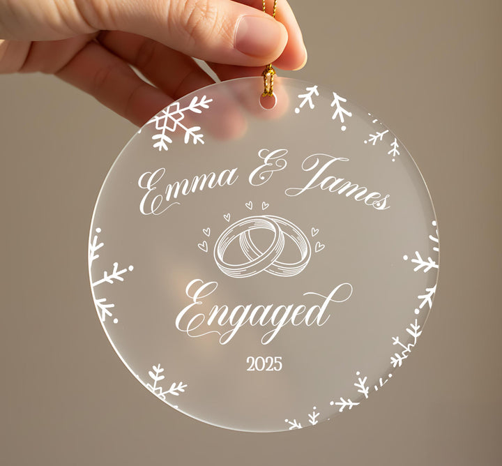 A hand holding a clear ornament with the text &quot;Emma & James Engaged&quot; and a graphic of a ring, surrounded by snowflakes.