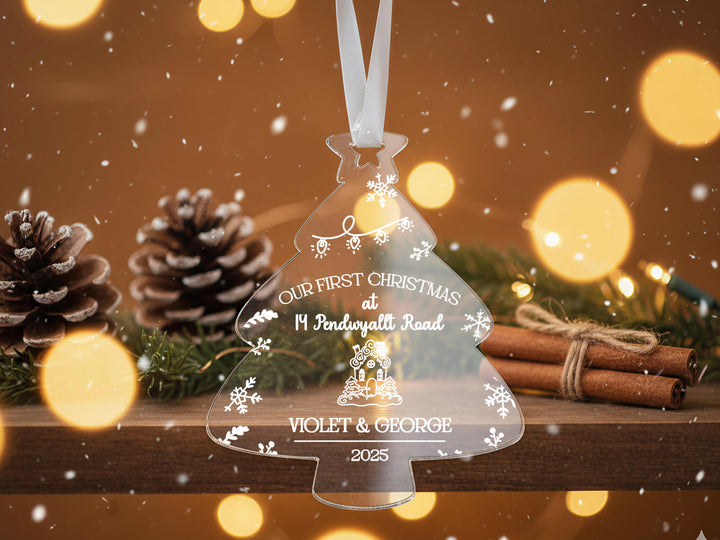 A clear Christmas ornament shaped like a tree hangs from a ribbon, surrounded by pine cones, cinnamon sticks, and twinkling lights, with the text &quot;Our first Christmas at 14 Pendewall Road&quot; and &quot; Violet & George 2020&quot; displayed on it.