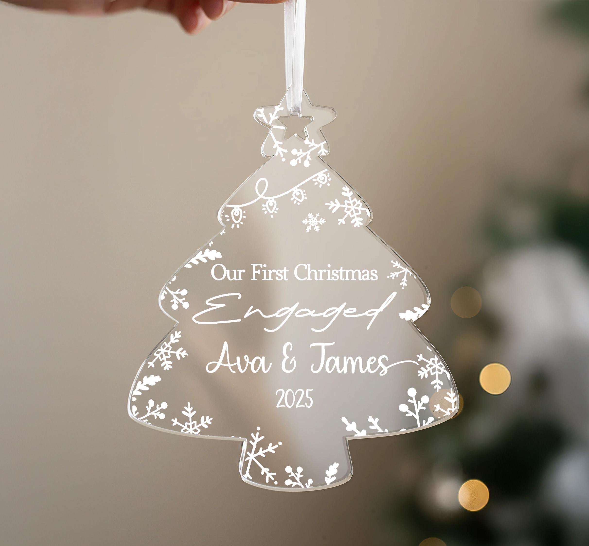 A hand holding a clear ornament shaped like a Christmas tree with the text "Our First Christmas Engaged Ava & James 2025" written on it.
