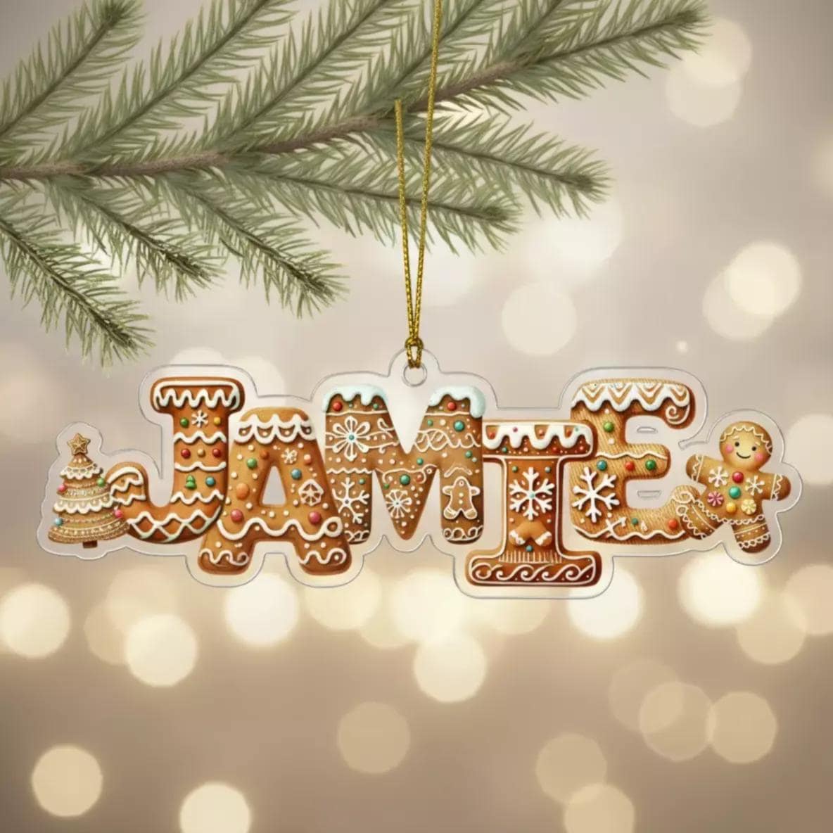 A clear ornament shaped like the name "JAMIE" made of gingerbread cookies, hanging from a branch with pine needles and a bokeh background.
