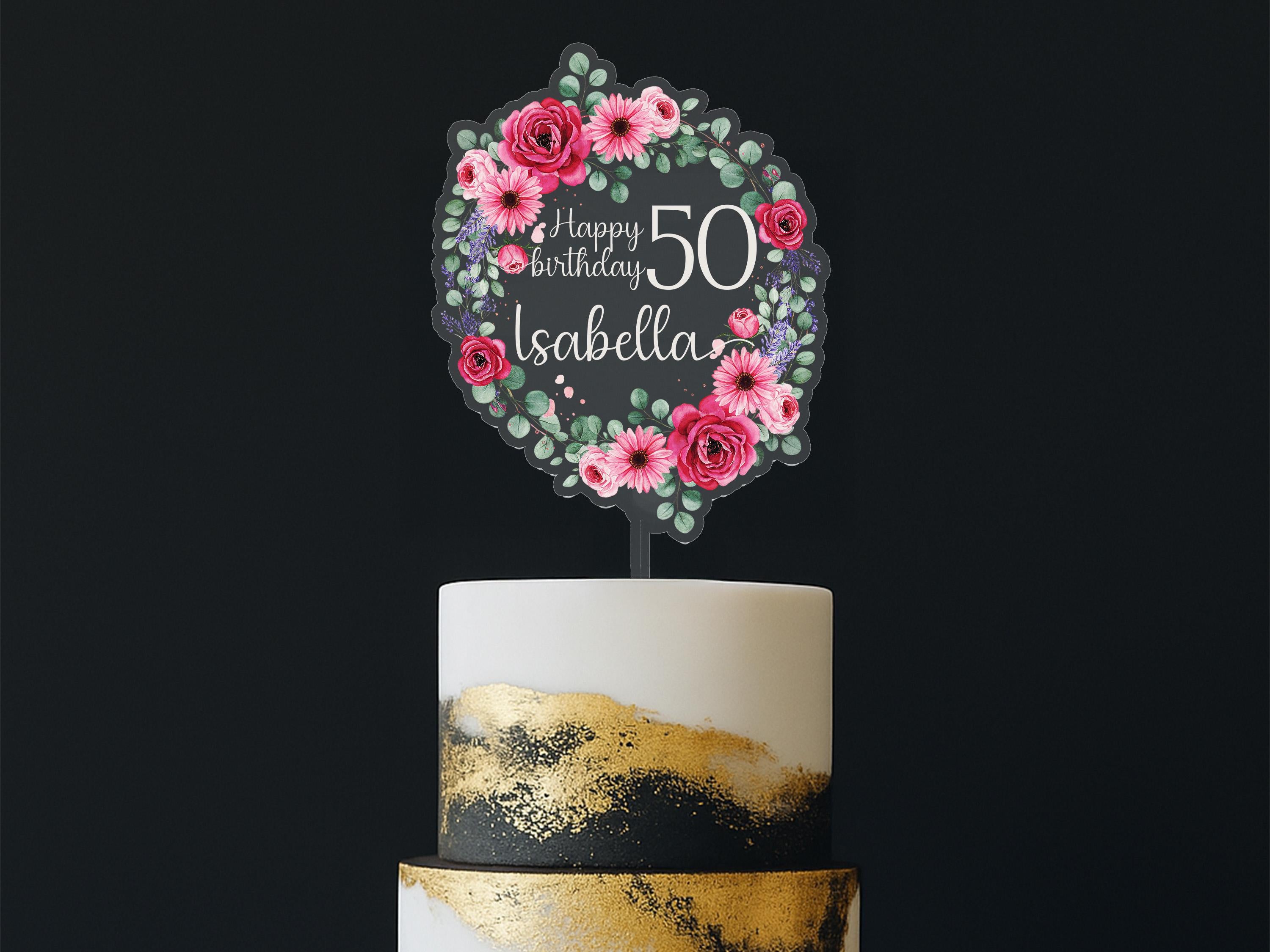 A birthday cake with a floral wreath-shaped topper that says "Happy Birthday Isabela" and has pink and red flowers surrounding it.