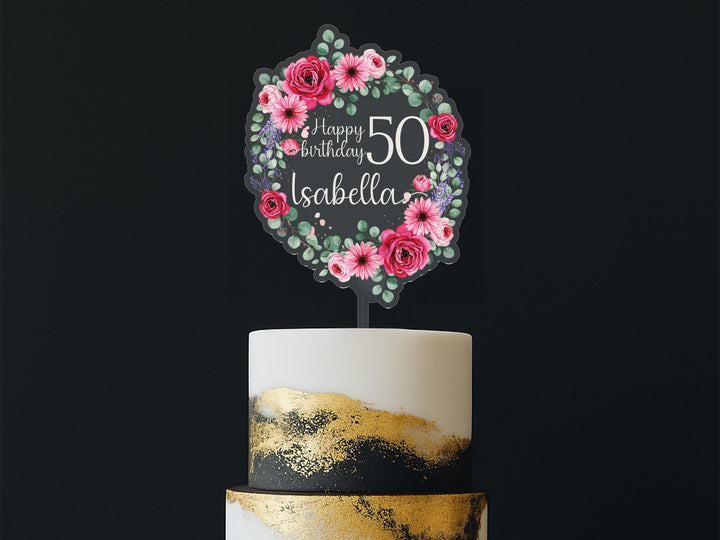 A birthday cake with a floral wreath-shaped topper that says &quot;Happy Birthday Isabela&quot; and has pink and red flowers surrounding it.