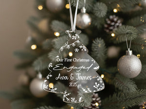 A clear Christmas ornament shaped like a tree hangs on a decorated tree, with the text "Our first Christmas Engaged Ava & James 2020" displayed on it.