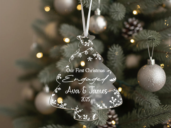 A clear Christmas ornament shaped like a tree hangs on a decorated tree, with the text &quot;Our first Christmas Engaged Ava & James 2020&quot; displayed on it.