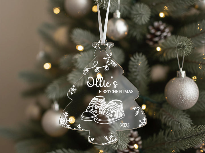 A clear Christmas ornament shaped like a tree hangs on a decorated tree, with a personalized message and a first- christmas date visible.