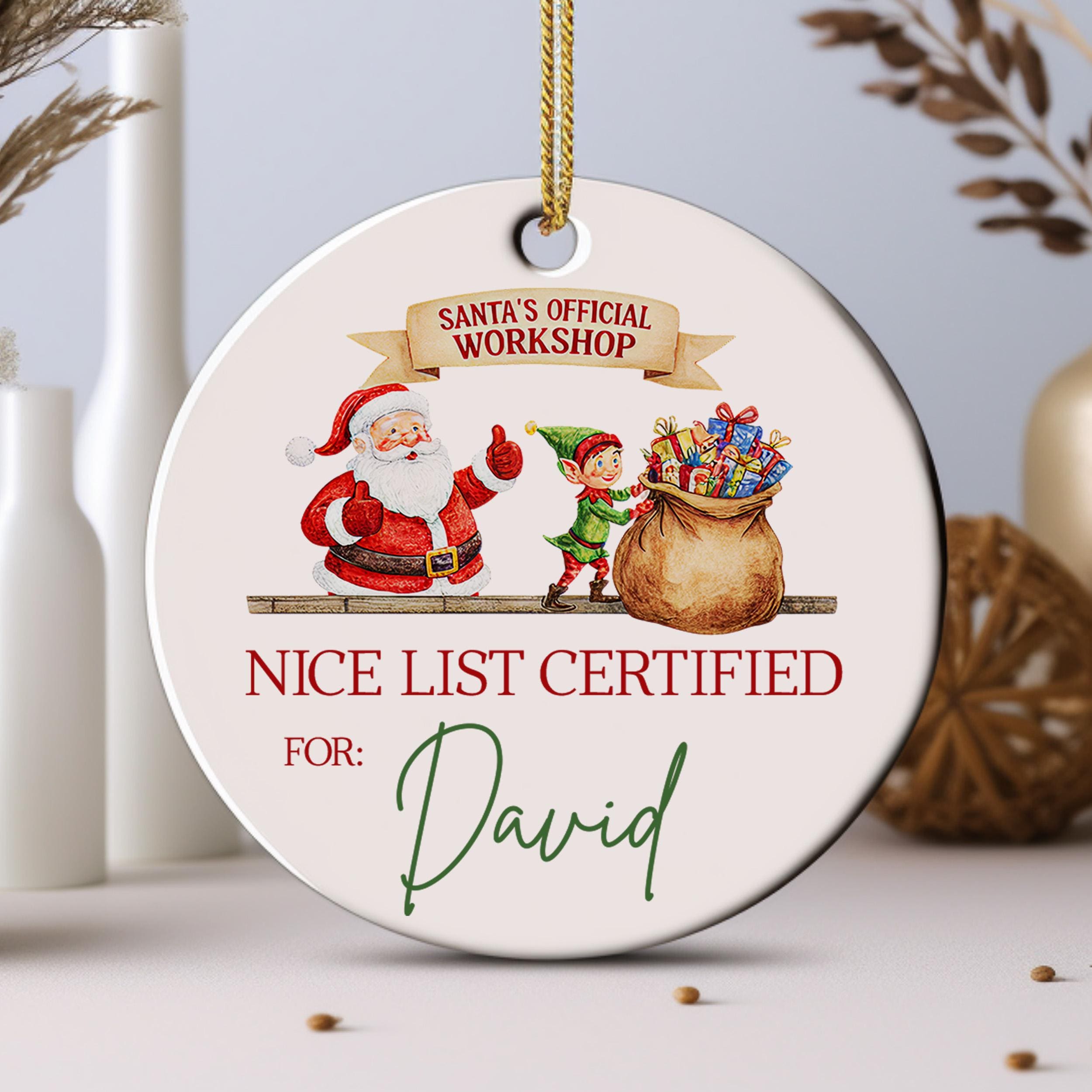 A white ornament with a Santa Claus and an elf, along with a bag of presents, hangs from a string. The ornament has text that reads "Santa's Official Workshop" and "NICE LIST CERTIFIED FOR: David".