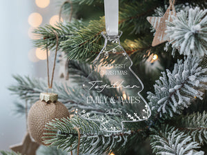 A clear Christmas ornament with the text "Together Emily & James" is hanging on a pine tree, surrounded by other ornaments and lights.