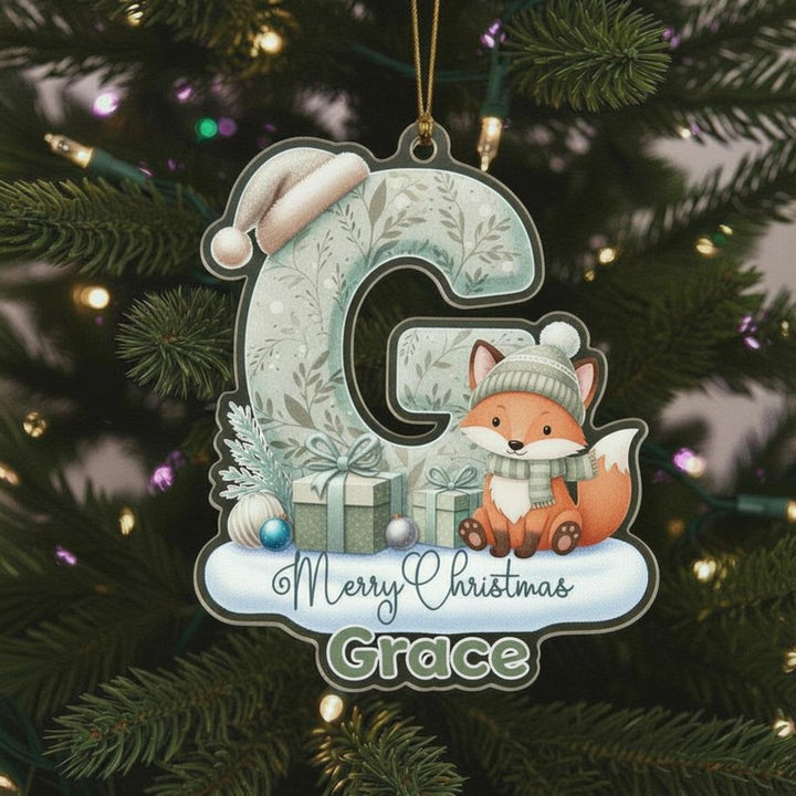 A Christmas ornament shaped like the letter &quot;G&quot; with a fox wearing a hat and scarf, sitting on a gift box, and surrounded by snow and Christmas decorations.
