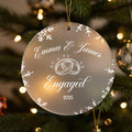 A clear ornament with a white design hangs on a Christmas tree, displaying the text "Emma & James Engaged" and the year "2025".