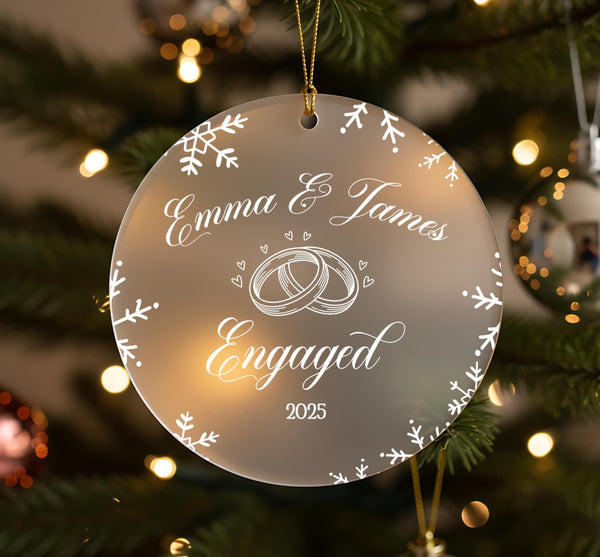 A clear ornament with a white design hangs on a Christmas tree, displaying the text &quot;Emma & James Engaged&quot; and the year &quot;2025&quot;.