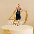 A woman in a black dress is standing on top of a white cake with the number 40 and the name "Andrea" written on it.