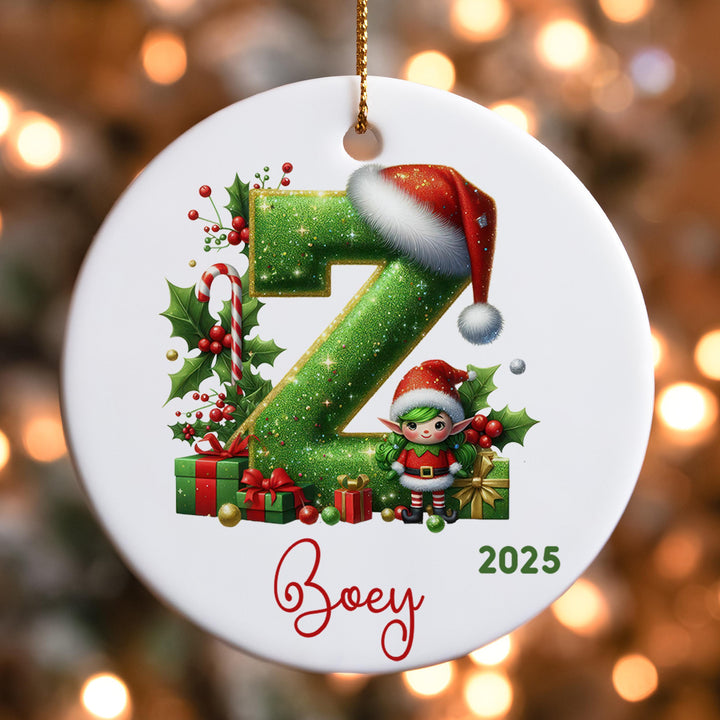 A white ornament with a green glittery letter &quot;Z&quot; and a small elf wearing a Santa hat, surrounded by Christmas decorations such as presents and holly leaves.