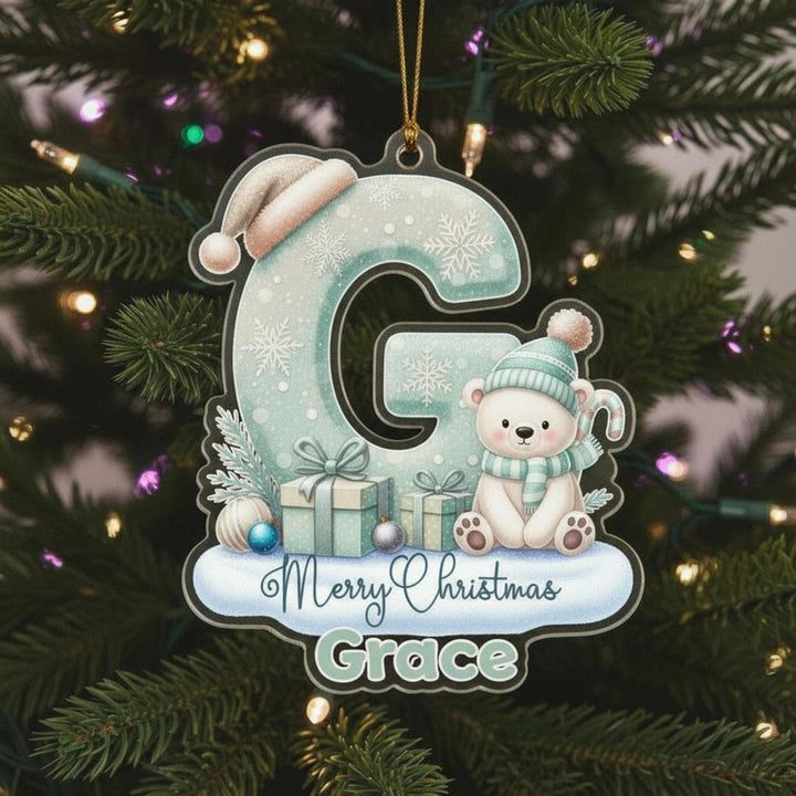 A Christmas ornament with a polar bear wearing a hat and scarf, sitting on a gift box, and surrounded by snowflakes and presents. The ornament is hanging on a Christmas tree with lights visible in the background.