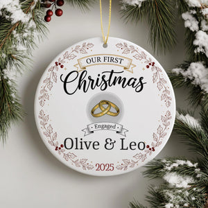 A white Christmas ornament with a gold ring and the text "Our first Christmas" and "Engaged Olive & Leo" printed on it, hanging on a pine tree branch.