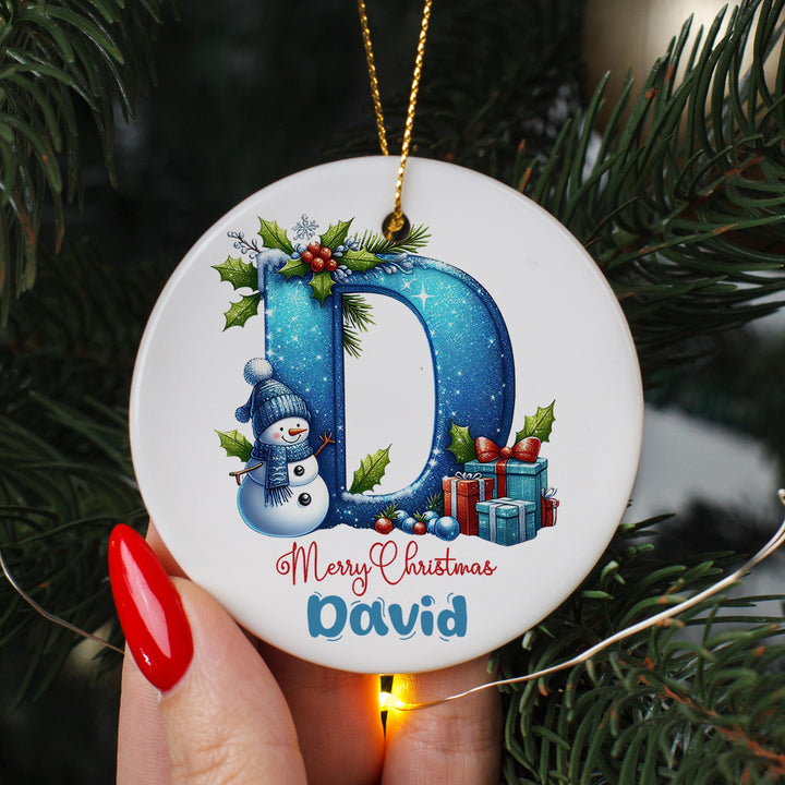 A hand holding a round ornament with a snowman and presents, surrounded by Christmas decorations and greenery.