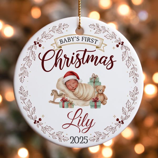 A white ornament with a baby and presents, surrounded by a decorative border and text, hangs against a blurred background with Christmas lights.