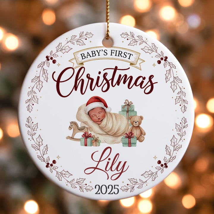 A white ornament with a baby and presents, surrounded by a decorative border and text, hangs against a blurred background with Christmas lights.