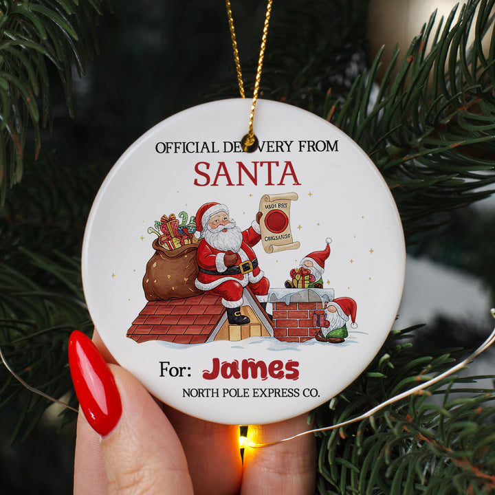 A person&#39;s hand holding a white ornament with a Santa Claus illustration and text, including &quot;Official Delivery From Santa&quot; and &quot;For: James North Pole Express Co.&quot;