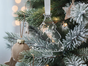 A clear ornament shaped like a Christmas tree with the names "Isabella & David" and the year "2025" written on it, hanging from a tree branch.