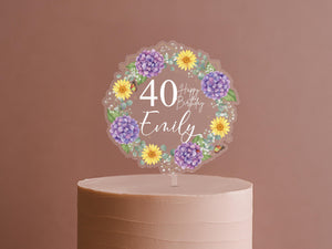 A cake topper with a wreath of purple and yellow flowers surrounding the number 40, along with the text "Happy Birthday Emily".