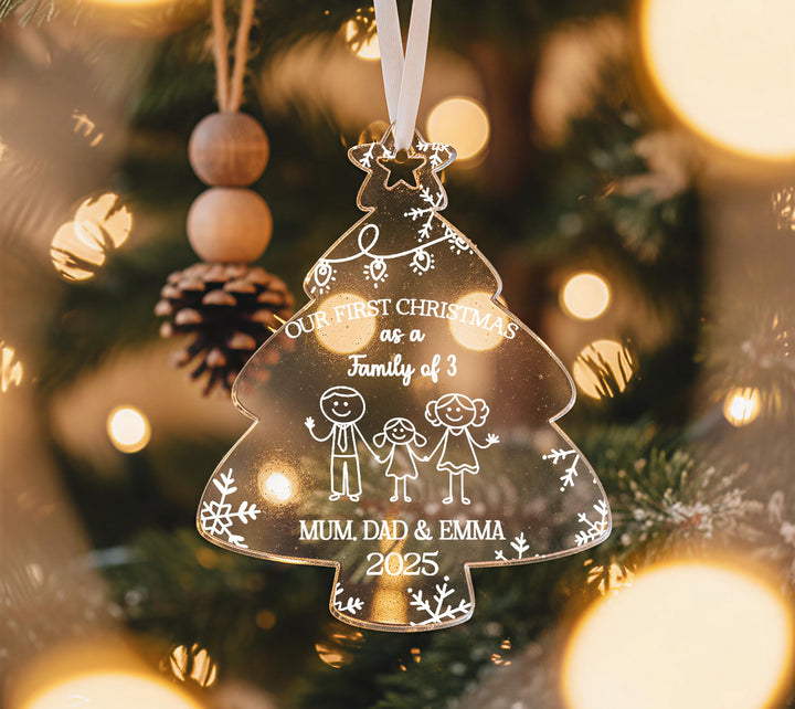A clear glass ornament shaped like a Christmas tree hangs on a tree, with a message about a family of three.