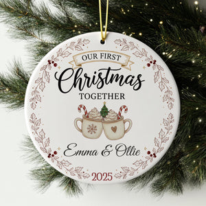 A white Christmas ornament with the text "Our first Christmas together" and an illustration of two mugs with hot chocolate and candy canes.