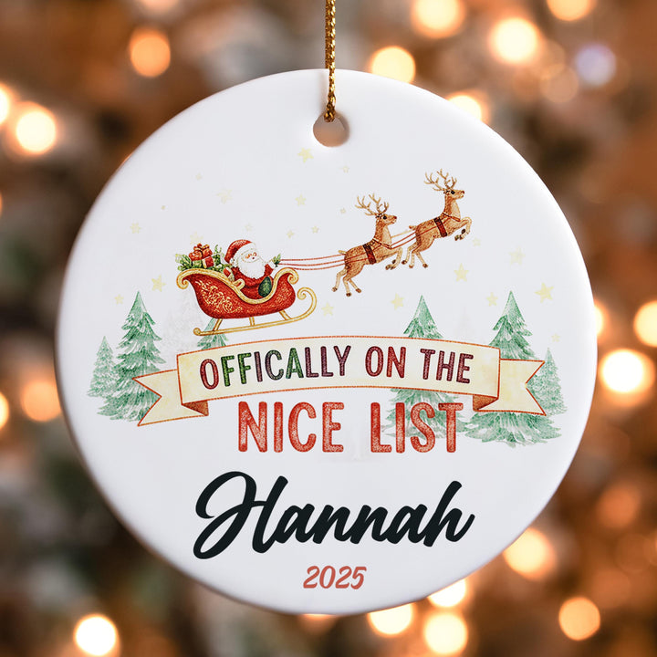 a white ornament with a Santa Claus sleigh in the background, along with a banner that says &quot; Officially on the NICE LIST Hannah&quot; and a year &quot;2025&quot; below it.