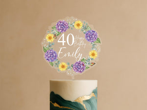 A birthday cake with a wreath-shaped topper that says "Happy Birthday Emily" surrounded by purple and yellow flowers.