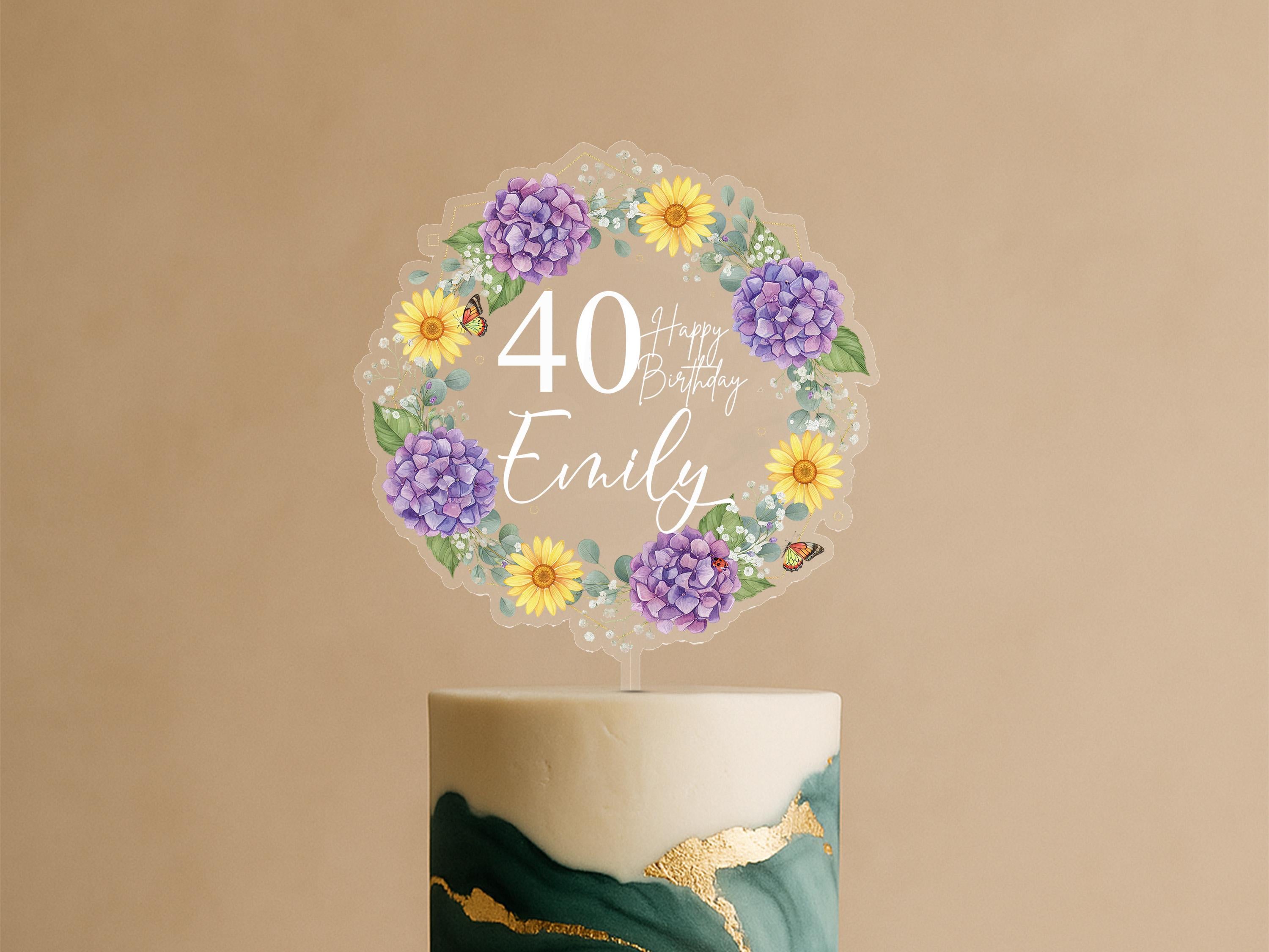A birthday cake with a wreath-shaped topper that says "Happy Birthday Emily" surrounded by purple and yellow flowers.