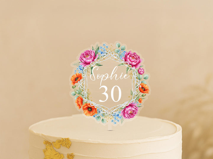 A white cake with a floral wreath-shaped topper that says &quot;Sophie 30&quot; on it.