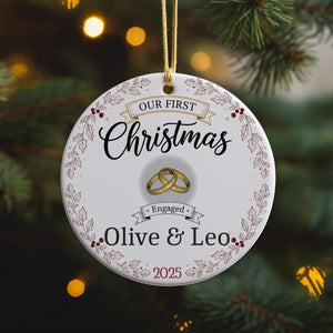 A white ornament with a gold ring and the text "Our first Christmas" and "Engaged Olive & Leo" printed on it, hanging on a Christmas tree.