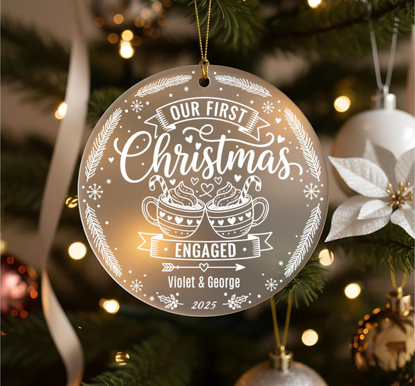Personalised first Christmas engaged ornament hanging on Christmas tree showing frosted acrylic design with hot chocolate mugs, hearts, snowflakes and festive details, engraved with couple names Violet and George 2025, surrounded by bronze baubles and pine branches